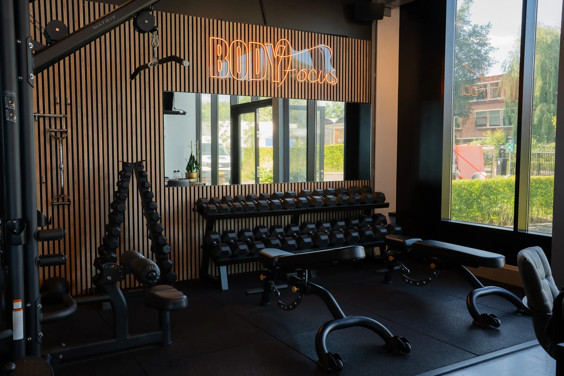 Dumbbellrek en trainingsbanken onder het oranje Body Focus neonlogo in de Open Gym