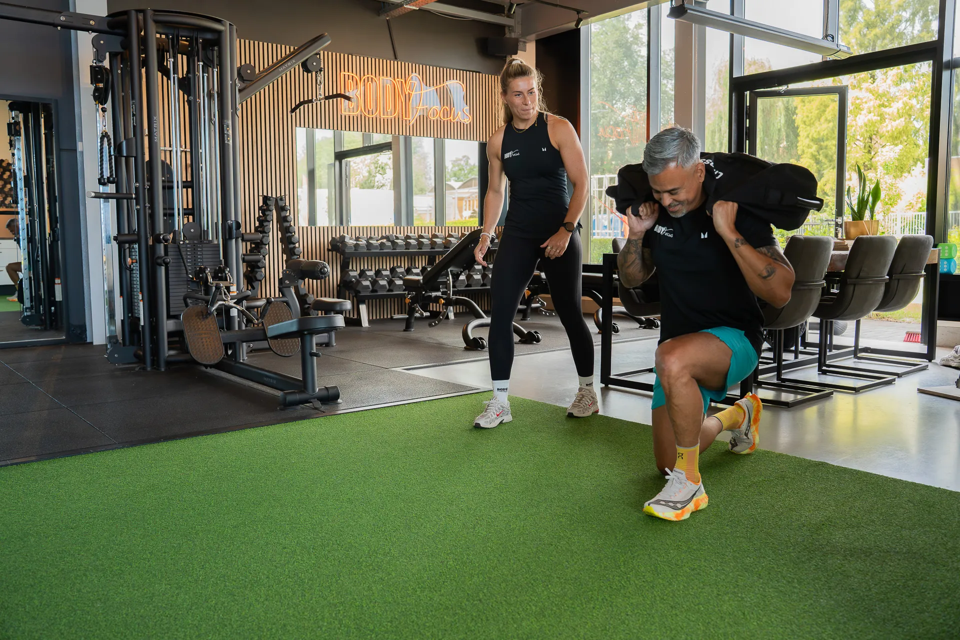 Trainer begeleidt member tijdens sandbag lunge op het kunstgras bij Body Focus